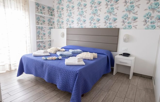 camera con letto matrimoniale con carta da parati floreale, ampia porta finestra e con teli spiaggia e asciugamani sui letti
