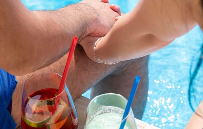 coppia si tiene per mano a bordo piscina con du cocktail tra loro