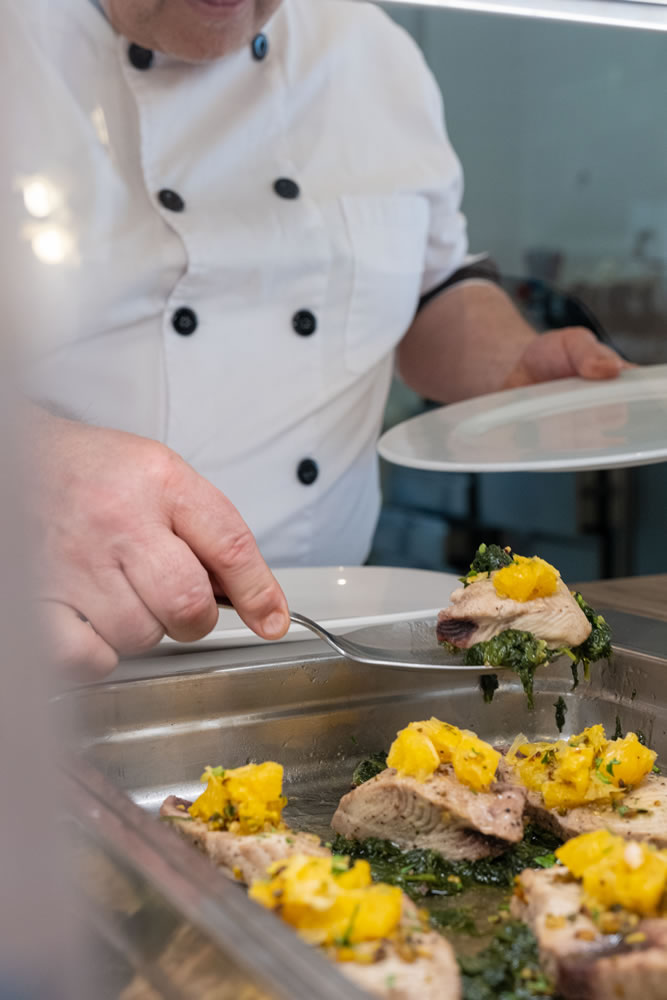 Chef raccoglie dalla teglia un cucchiaio di pesce al forno con patate e bieta saltata
